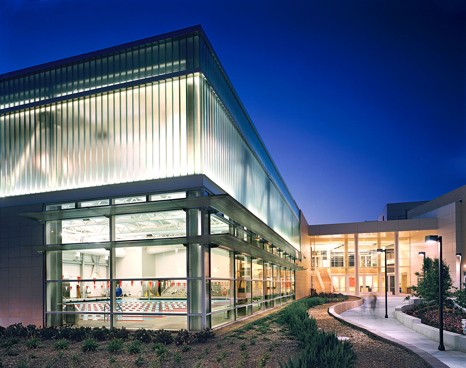 corner view of exterior elevation at night showing clear and frosted floor to roof panels over an indoor swimming pool and entry way and landscaping